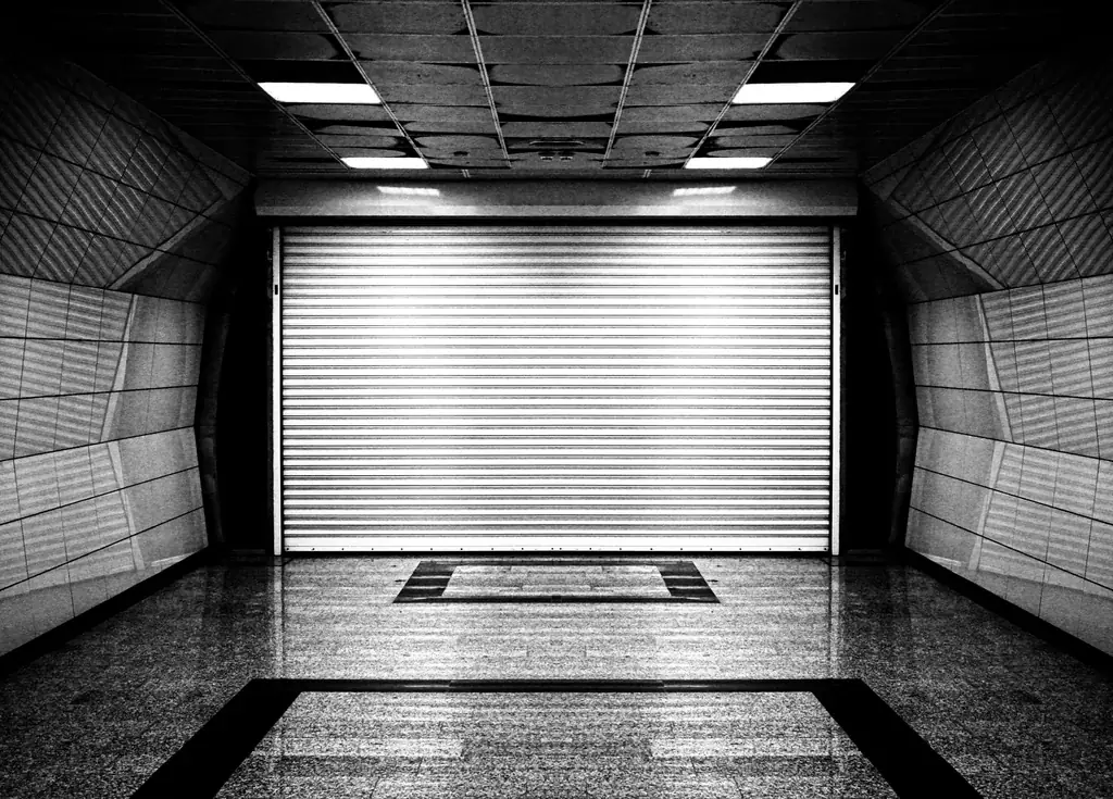 Black and white photo of a closed roll-up door at the end of a tiled corridor