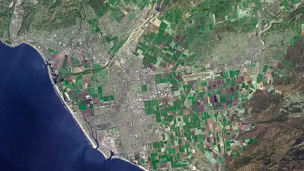 Satellite view of a coastal plain with a river mouth, farm fields and a city against the hills